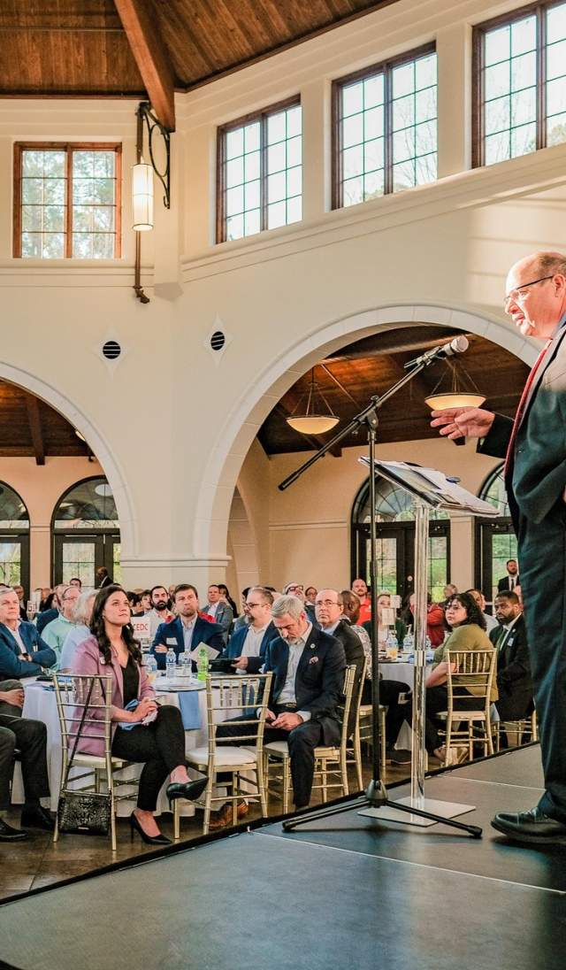 A professional speaker addresses a full room of attendees seated at banquet tables inside a bright, elegant event space at Cape Fear Botanical Garden in Fayetteville, North Carolina.