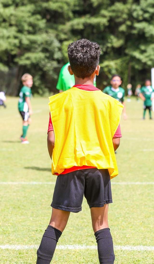 A child wearing a yellow practice vest stands on a grassy soccer field and looks toward a youth match in progress, with teammates, goals, and tree-lined surroundings visible ahead.