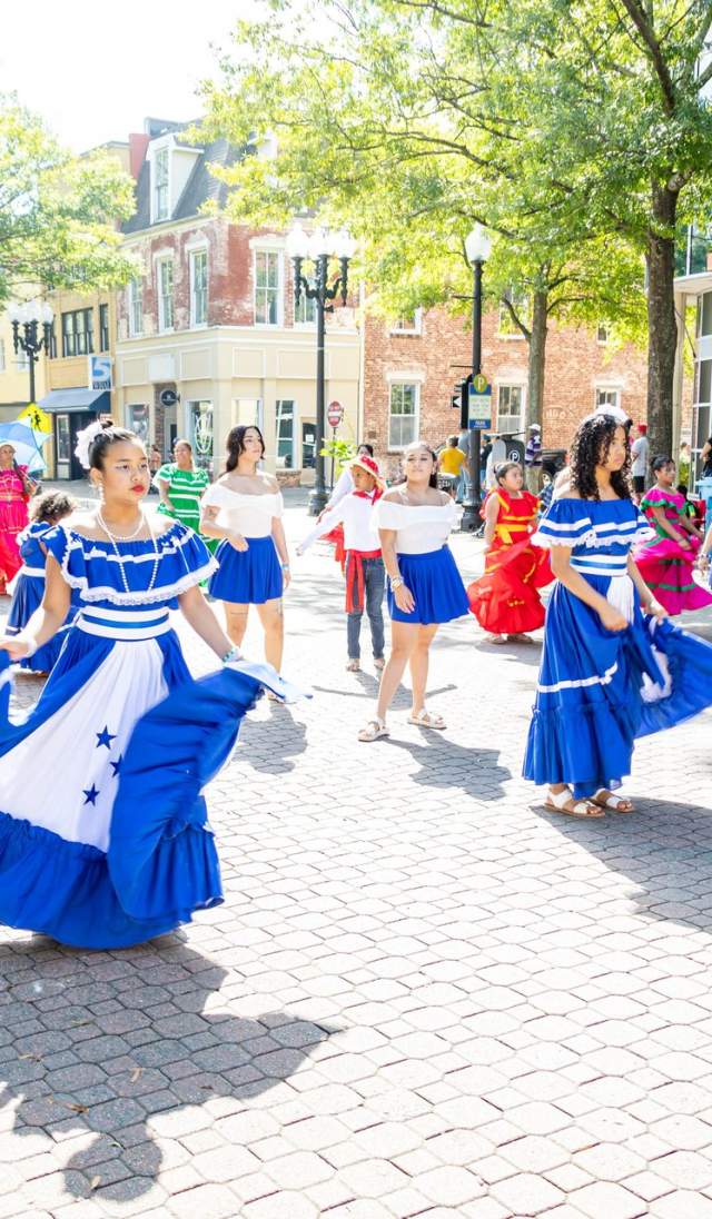 Dancers in blue and white dresses perform in downtown Fayetteville during a daytime cultural festival, with spectators lining the brick street.