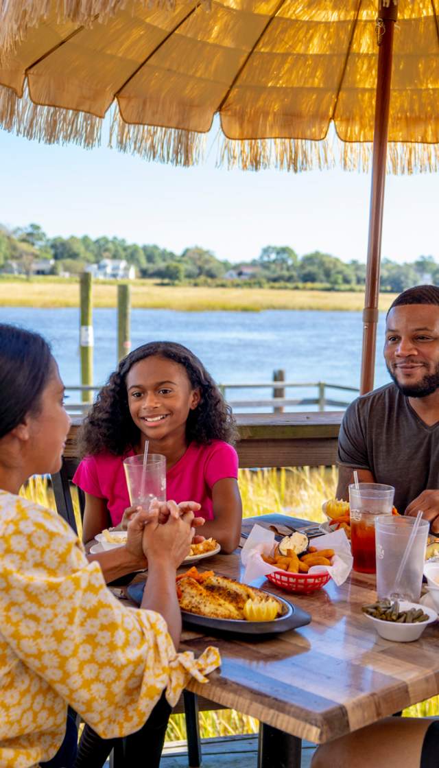 a family of four eating on an outside patio with a view of a shrimp boat and river in the background
