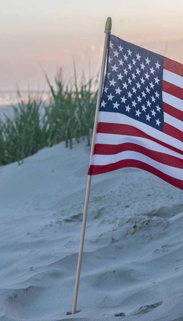 a small American Flag placed in the sand on a beach at sunset