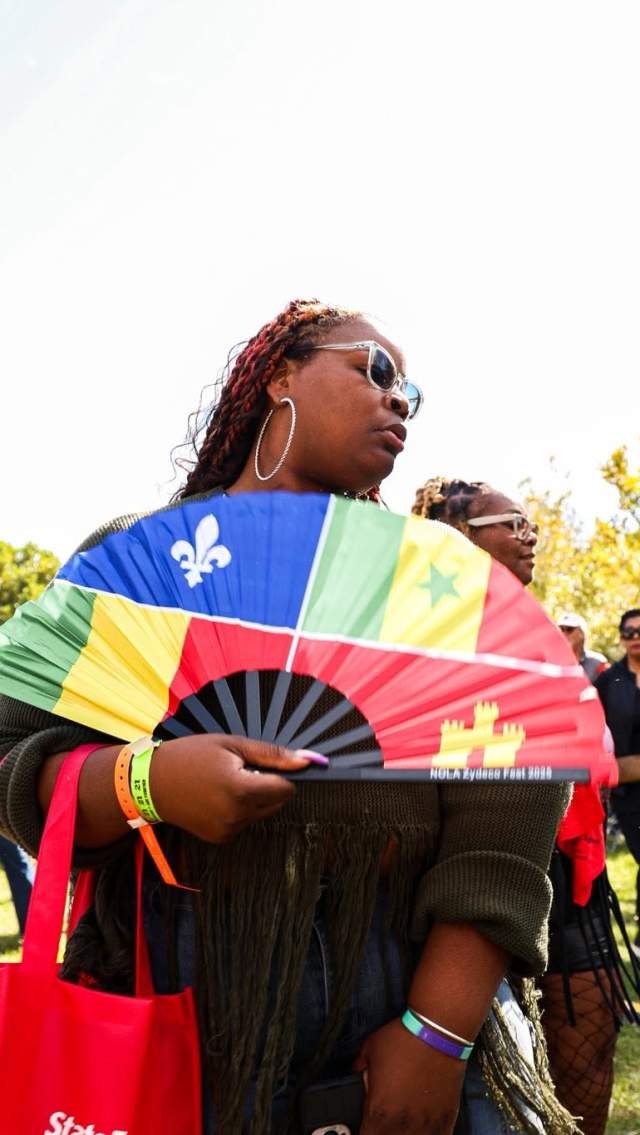 Festivalgoers dance on the lawn at NOLA Zydeco Fest, including a woman holding a colorful fan featuring Louisiana and West African flags.
