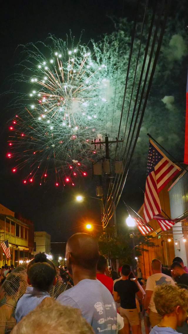 Crowds fill the streets of Olde Towne Slidell during a Fourth of July celebration as fireworks burst overhead. American flags line the storefronts while families gather beneath the night sky to watch the colorful display.