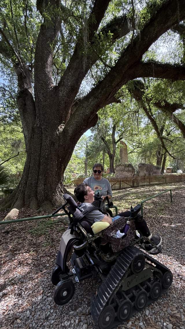 Christy Paulsell accompanies her daughter, PJ, in an all-terrain TrackChair beneath towering live oaks at Fontainebleau State Park, with the historic sugar mill ruins in the background.