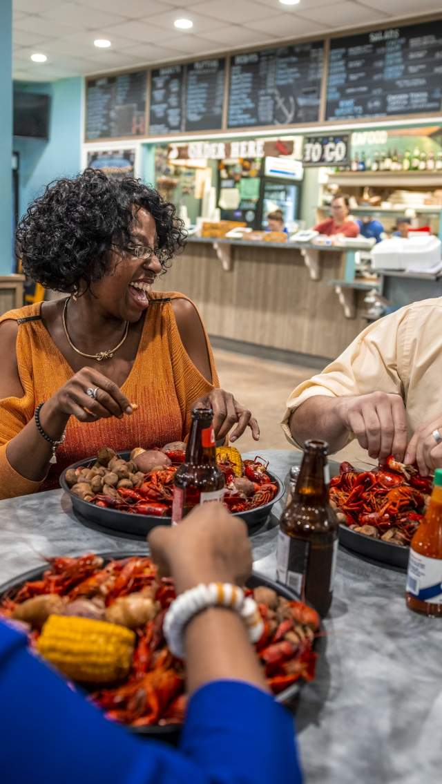 At Mandeville Seafood, two women and a man dig into trays of boiled crawfish with all the fixings. The wall menu and order counter is visible behind.