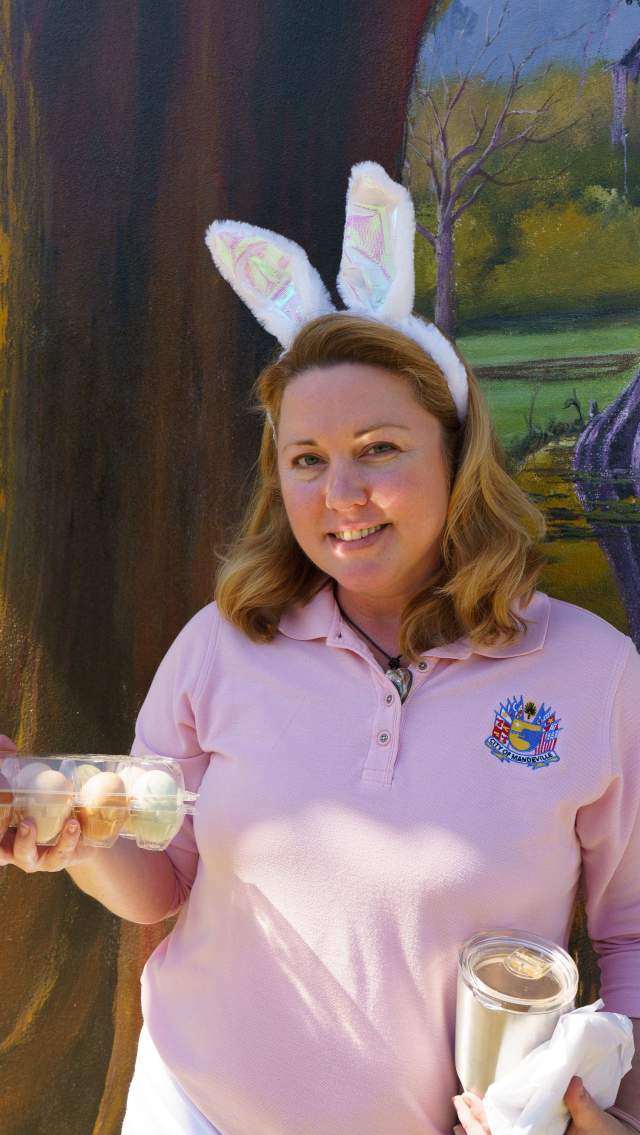 A woman shopping at Mandeville Trailhead Easter Market wears bunny ears and holds a carton of eggs and a jar. There is a mural on the wall behind.