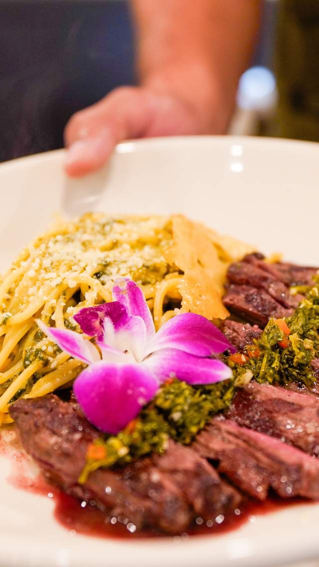 A plate of sliced meat and pasta with a hot pink edible flower on top