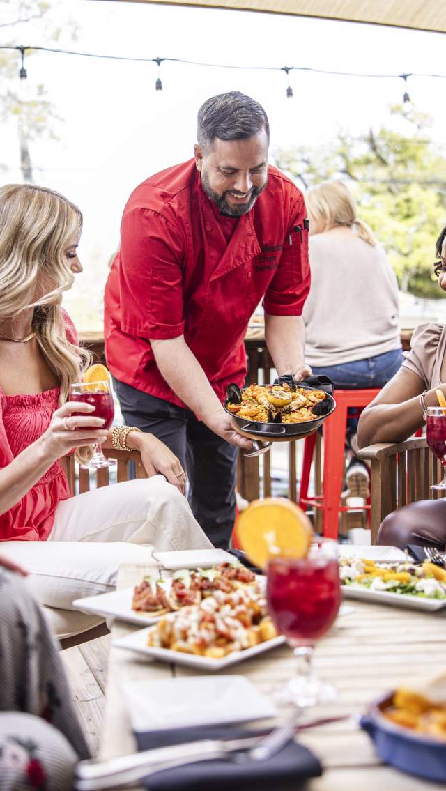 Three women are seated around a low table at Valencia Restaurant's outdoor patio as the chef sets a dish on the table, which is laid with various plates and drinks.