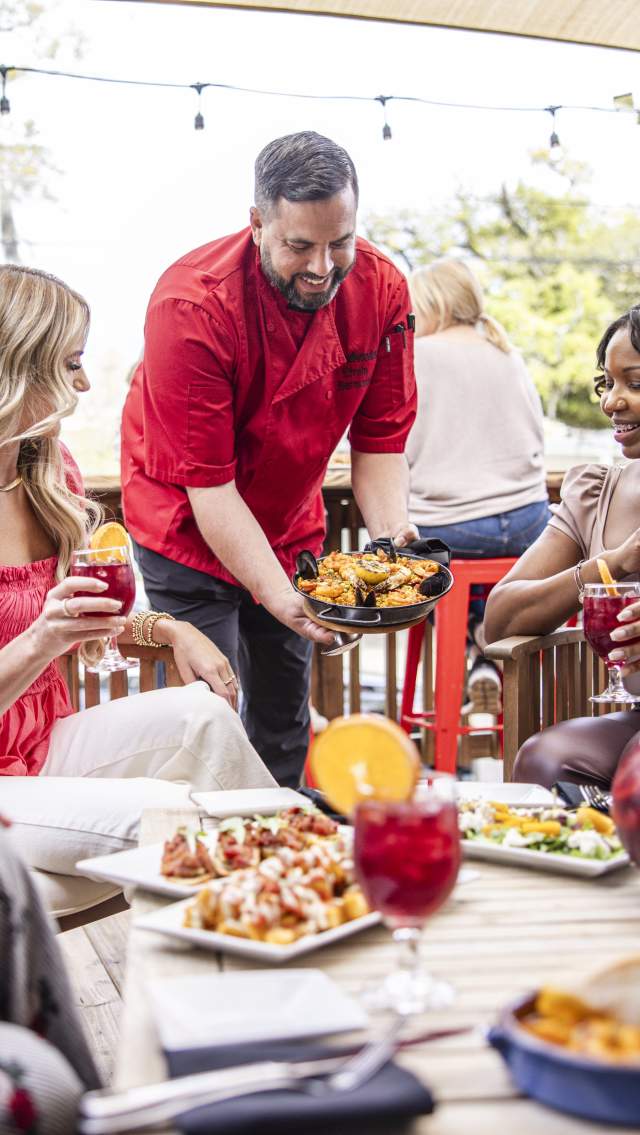 Three women are seated around a low table at Valencia Restaurant's outdoor patio as the chef sets a dish on the table, which is laid with various plates and drinks.