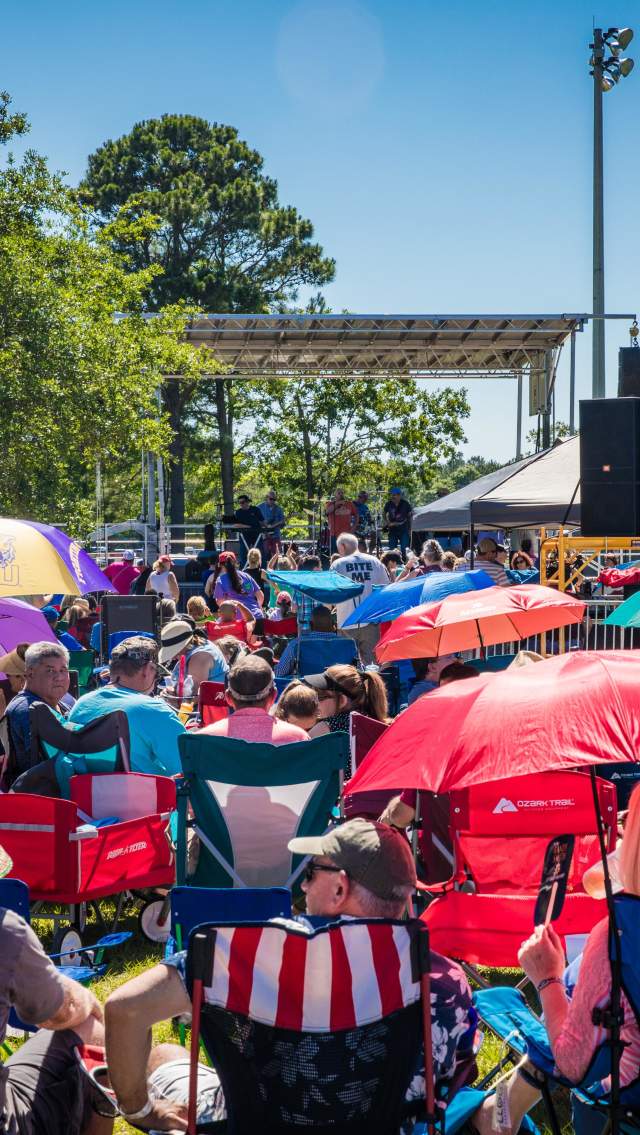 A large crowd sits in lawn chairs and under colorful umbrellas facing a stage during the Slidell All-You-Can-Eat Crawfish Cook-off on a sunny day.