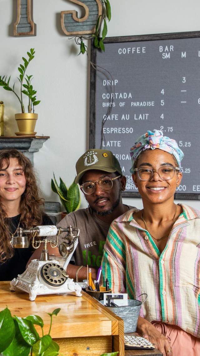 Staff members and the co-owners stand behind the counter at Roots Plants+Coffee in Olde Towne Slidell, with coffee beans, houseplants and the café menu in the background.