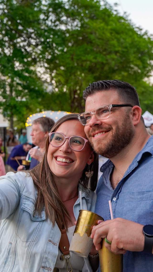 A couple smiles and takes a selfie while holding drinks during the Girod Street Stroll in Mandeville, with a lively crowd and vendor tents in the background.