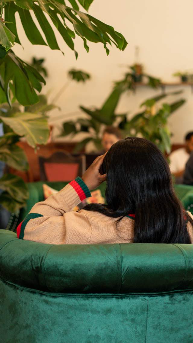 Customer seated in a green velvet chair inside Roots Plants & Coffee in Slidell, surrounded by large tropical plants and shelves of potted greenery, while other guests gather in the bright café background.