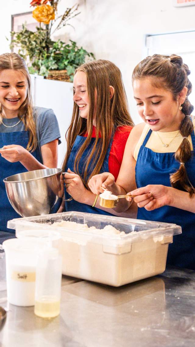 Four teen girls in blue aprons and a cooking instructor in a white smock stand at a kitchen prep table measuring cups of flour from a large, rectangular tub.