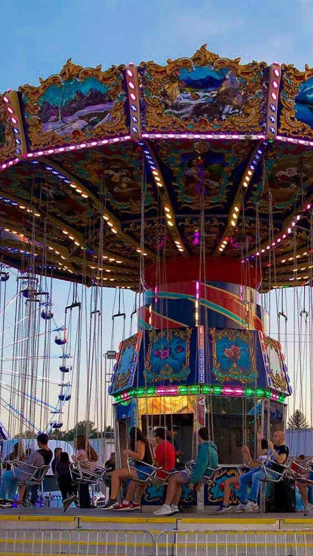 Festivalgoers sit aboard a brightly lit swing ride with a Ferris wheel in the background at Slidell Food & Fun Fest.