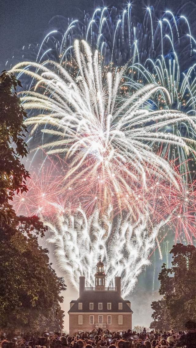 Fireworks illuminate the night sky above a historic building, surrounded by a crowd of spectators. Lush trees frame the scene, enhancing the festive atmosphere.
