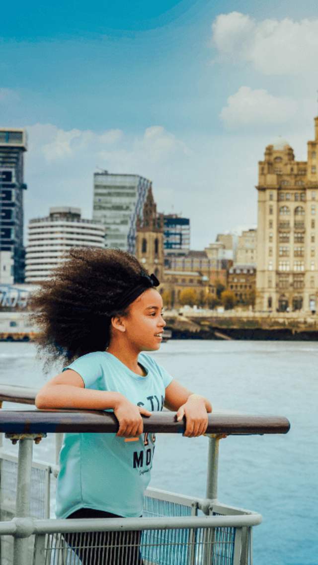 A young child on the Mersey Ferry. They are standing on the end next to the railings looking out over the river. In the background is the Three Graces, the royal Liver Building, Cunard Building and the Port Of Liverpool building.