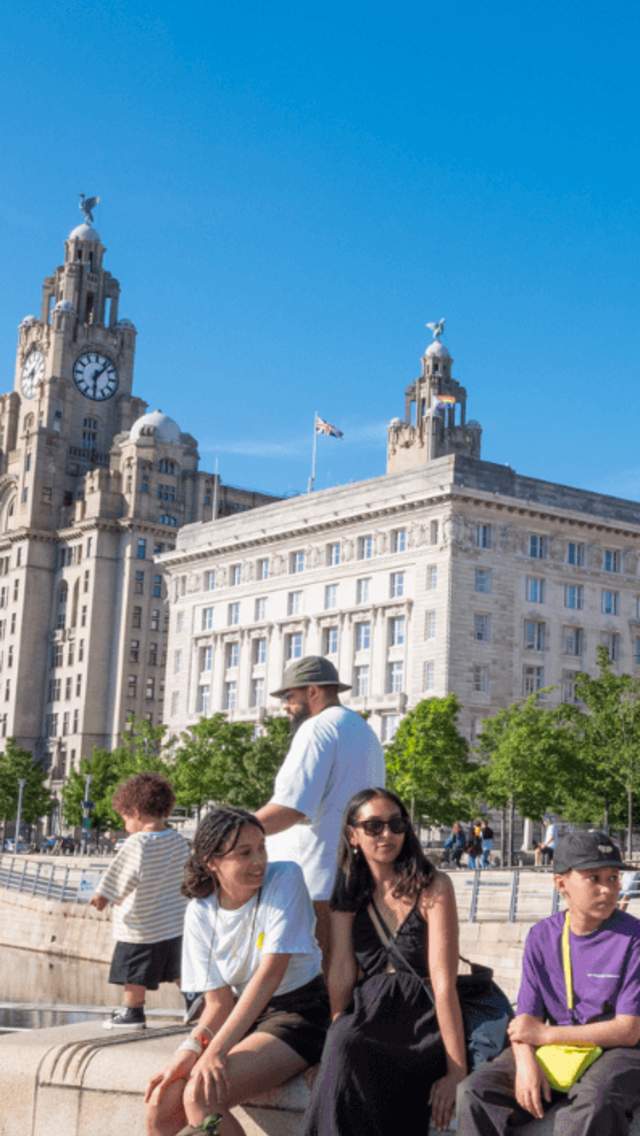 Liverpool Pier Head in the sunshine with a family sitting on a wall. A small child is holding a balloon in the shape of a rainbow.
