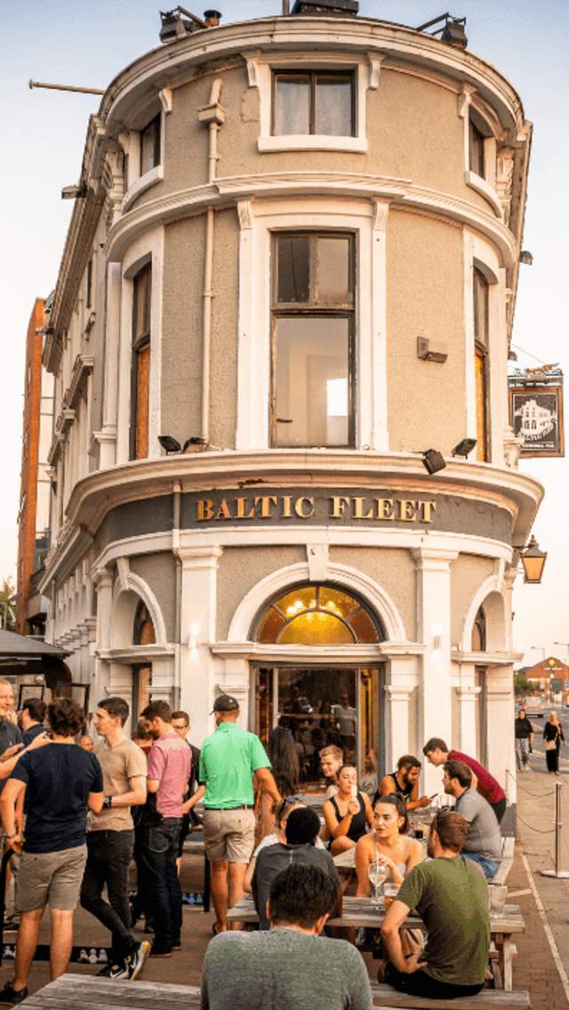 A summer evening outside the Baltic Fleet Pub. The narrow stand-alone pub building is centre of the shot, with the dock warehouse buildings to the right and modern apartments to the left. People are dressed in summer clothes sitting at wooden picnic benches.  i
