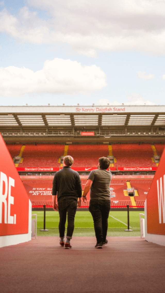 Two people are walking out of the players tunnel at Liverpool's stadium, Anfield. Either side of them are walls in red which have the words 'We are Liverpool. This means more'. In front of the two figures you can see the stand on the other side of the pitch and blue sky with few clouds.