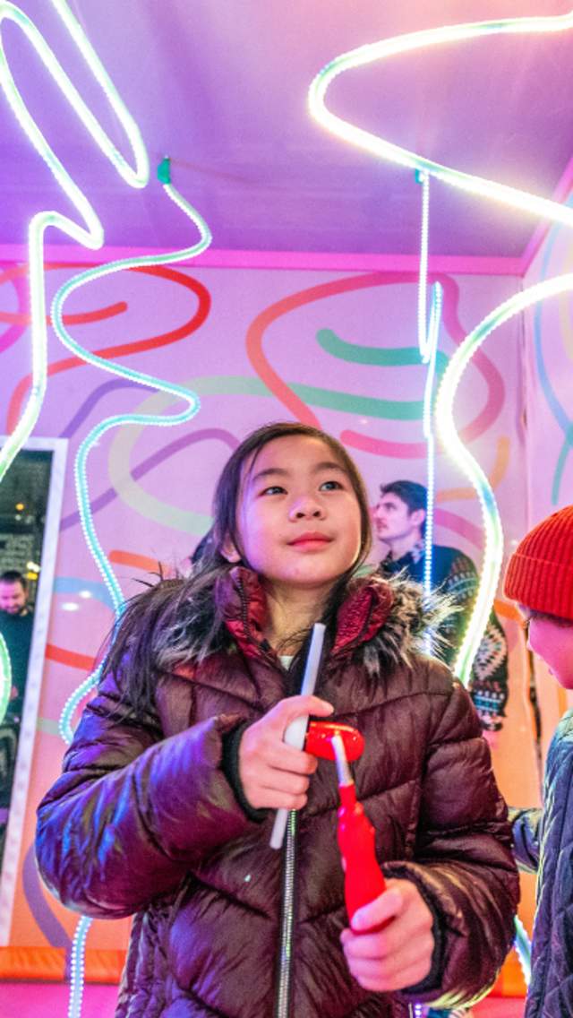 Inside a colourful pink box. There are ribbon shaped LED lights hanging from the ceiling. Children are smiling looking at the lights and an adult is watching the children smiling.