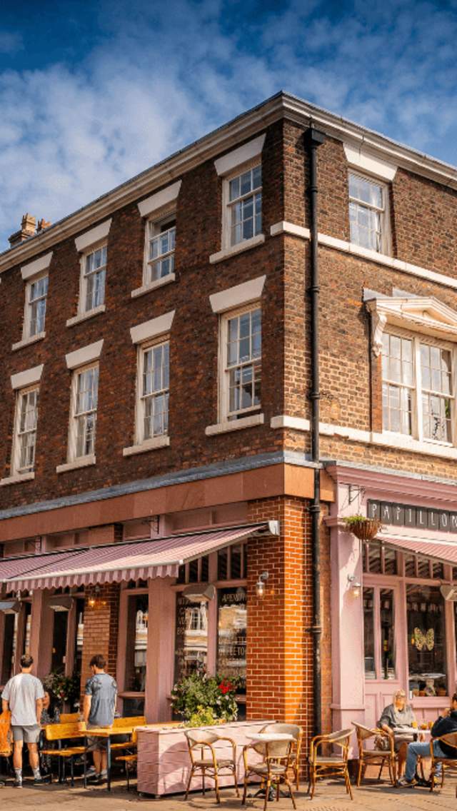A corner shot of a Georgian Town House style building with sash windows. The ground level of the building has been turned into a cafe, painted a complimentary light pink with awnings and outdoor seating. The sky is blue and it looks like a warm summer day.