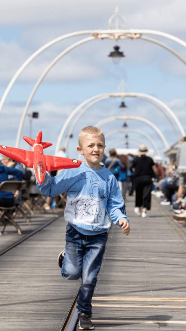 A young boy running along the Southport Traditional Pier. They are holding a blow up aeroplane