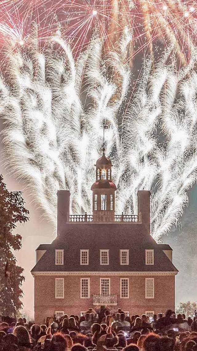 Fireworks illuminate the night sky above a historic building, surrounded by a crowd of spectators. Lush trees frame the scene, enhancing the festive atmosphere.