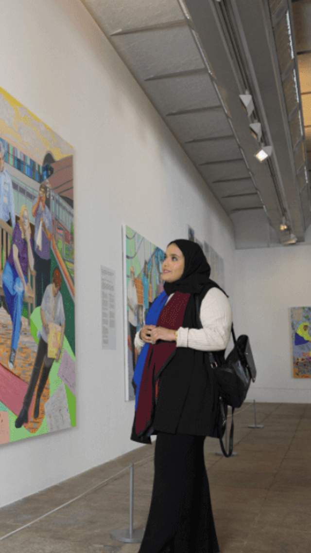 A lady wearing long black clothes looks at a large colourful and lifelike painting in a gallery. The gallery has white walls and grey metal supports.