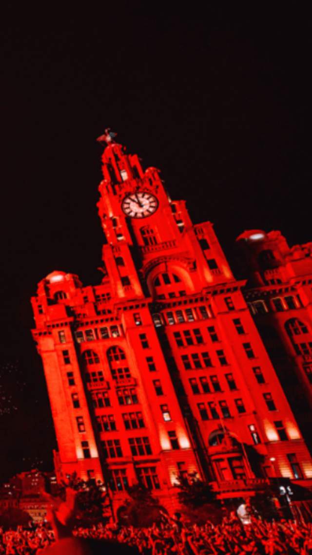 Liverpool Pier Head in the dark with the Liver Building bathed in red. To the left is a stage with fireworks on the roof.
