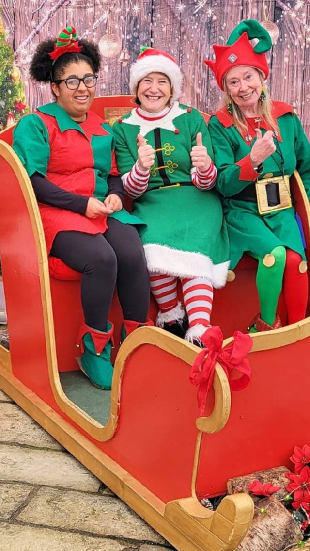 Three women dressed as Christmas characters sitting in a red sleigh