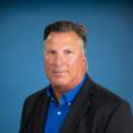 man with gray hair poses in black sports coat or suit and blue shirt