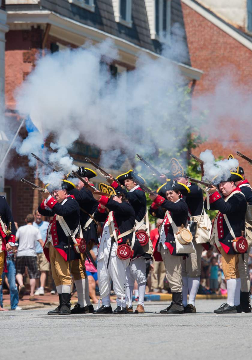 A group of historical reenactors in colonial military uniforms fires muskets, creating clouds of smoke during a lively outdoor event.