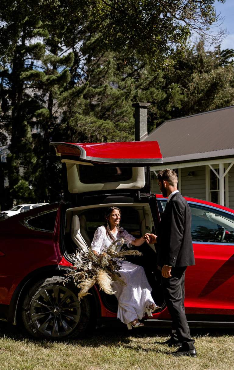 A couple at their wedding step out of a red Tesla vehicle