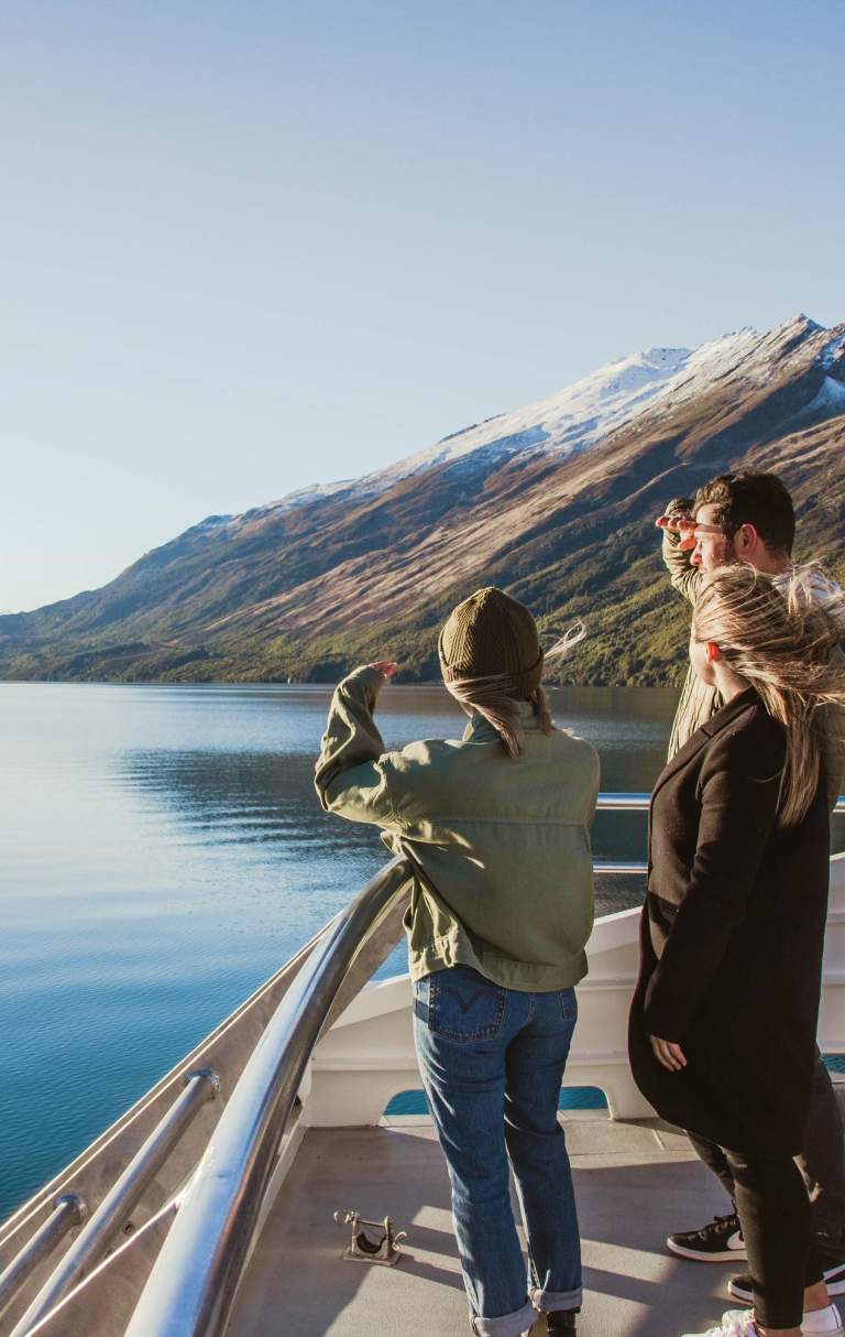 Three people standing on the deck of a boat, gazing out at the calm blue waters of a lake surrounded by mountains. The landscape features snow-capped peaks and lush greenery under a clear blue sky, creating a serene and picturesque setting