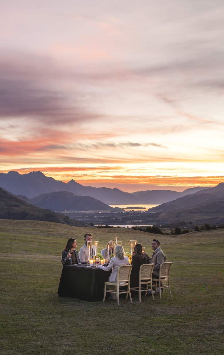 A group of corporate people dining outdoors beside a softly lit marquee, overlooking Queenstown’s mountains and Lake Whakatipu at sunset.