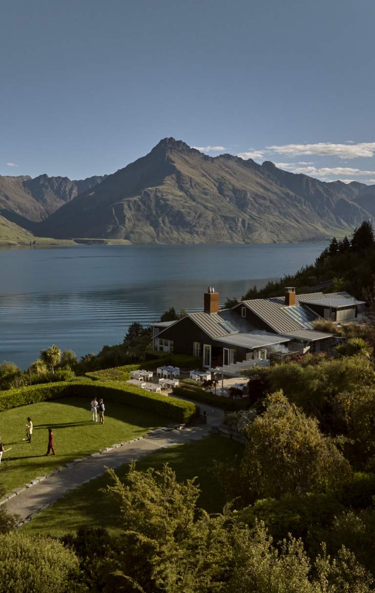 A drone shot of a luxury lodge surrounded by bush, on the shore of Lake Whakatipu with mountains in the background