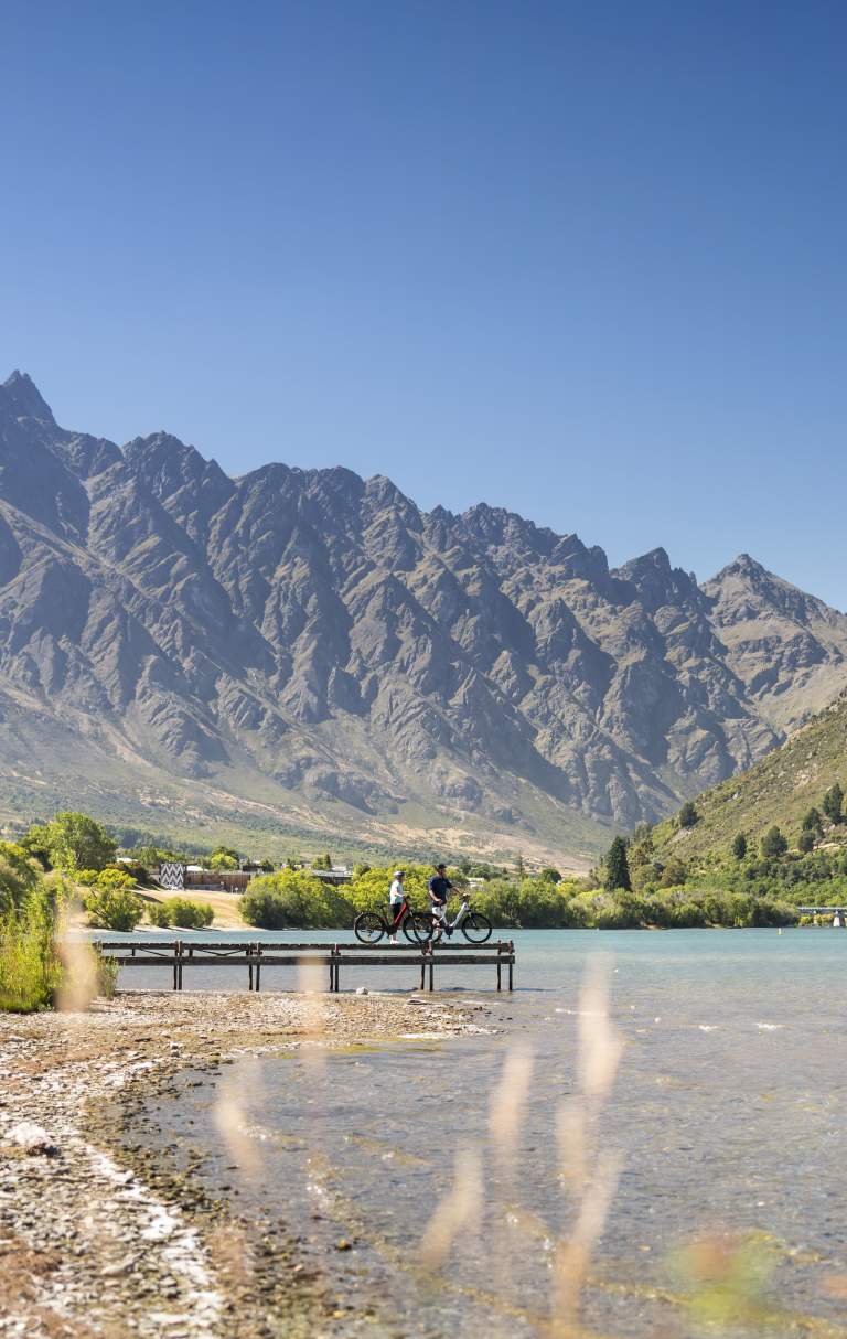 Two people having a rest on a pier looking out over the lake and Remarkables mountains while biking the Frankton Trail