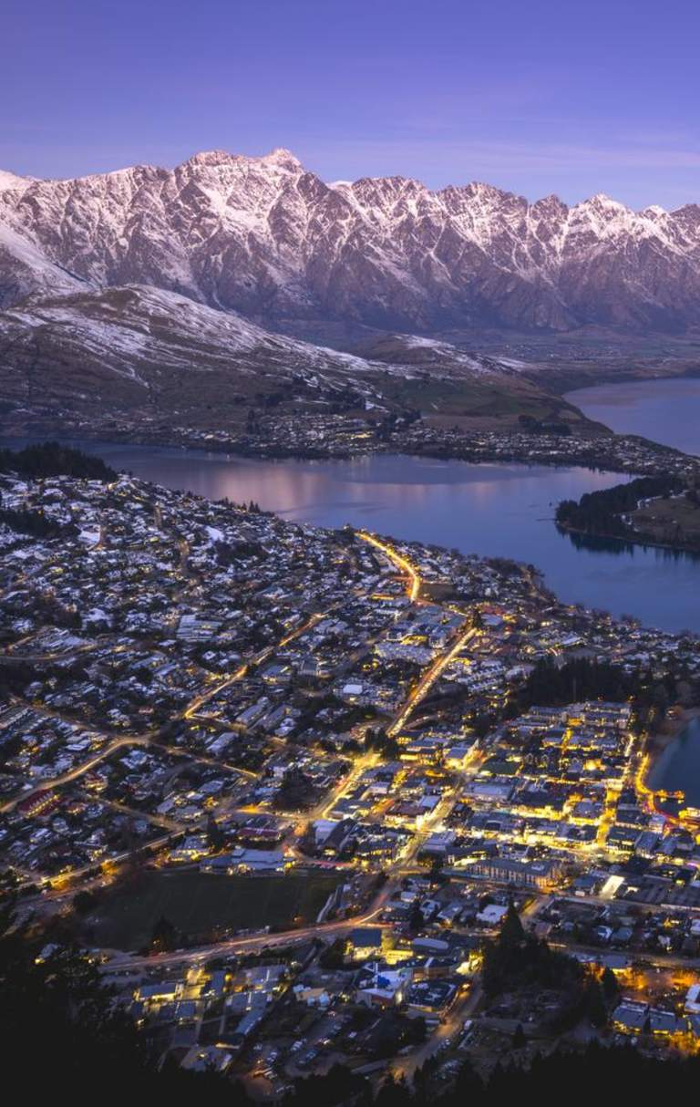 Aerial shot of Queenstown on a winters evening with town street lights glowing and snow capped mountains in the background