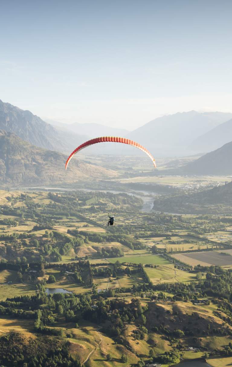 Person paragliding over a green valley with the Remarkables mountain range in the background