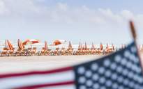 In focus is the background of the photo, which shows a row of orange and white beach chairs and umbrellas on the beach. An American flag is in the foreground, blurred.