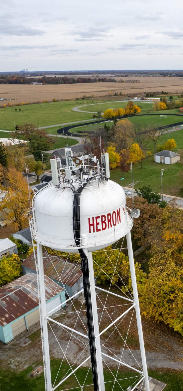 An aerial view of a town. A white water tower that says Hebron is in the foreground. A church, baseball diamond, and cornfields are in the background.
