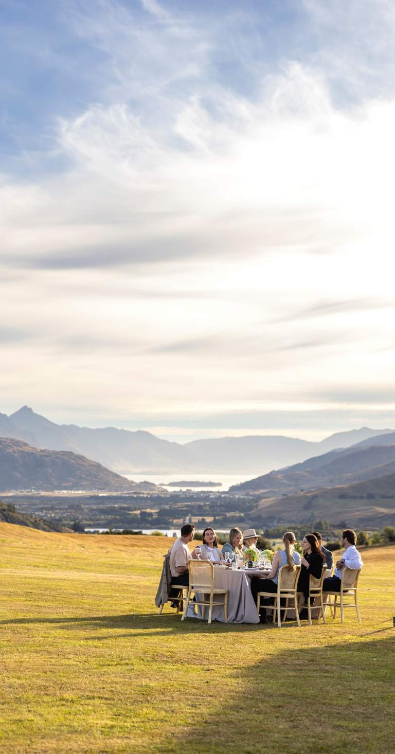 A group of people attending a business event function sitting around a table for lunch with large views of mountains, lake and basin surrounding them. There is a white tent next to them.
