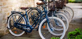 A row of blue bicycles with white tyres and brown wicker baskets stand by a wall at Calcot & Spa