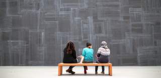 Photograph showing three people sat on a bench, taking a break as they visit an exhibition. The wall behind them is a slate grey with white lines