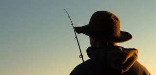 Photograph showing a person fishing from behind, with the dusk light only just illuminating them.