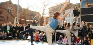 Three dancers stand on one leg in Trinity Square with a crowd surrounding them