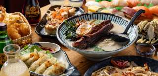 Spread of Japanese cuisine on wooden table including big bowl of ramen, dumplings, and sushi