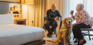 A couple relaxes in a modern hotel room at the DoubleTree by Hilton in Hull, sitting with a trained assistance dog beside them.