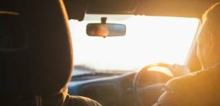 Photograph showing a man driving a car, as viewed from the back seats. The light is of the evening's golden hour.